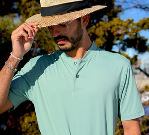 A man in a beryl green maelreg everyday Jersey Henley Shirt and straw hat, standing outdoors and adjusting his hat.